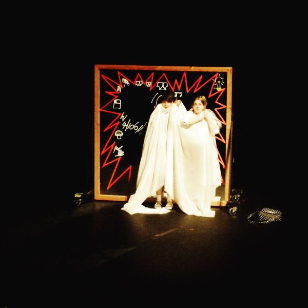black background on a stage. A square mirror with red and white. patternes and writing on it. The mirror has a light brown frame around it. Two Women are standing in front of the mirror . with a white sheet over them